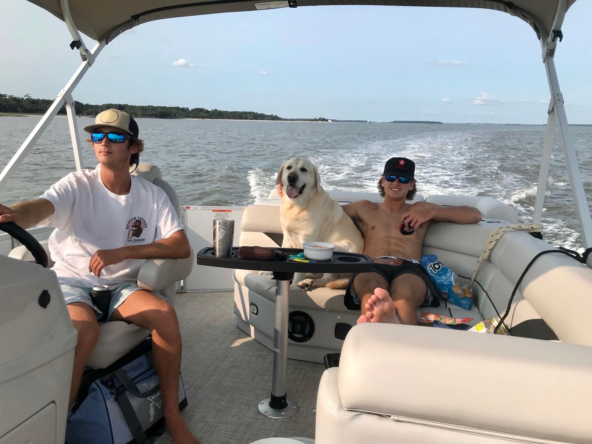 Two young men and a smiling yellow Labrador retriever relaxing on a pontoon boat cruising on a calm lake under a clear sky, snacks and drinks on a small center table and the boat wake trailing behind.