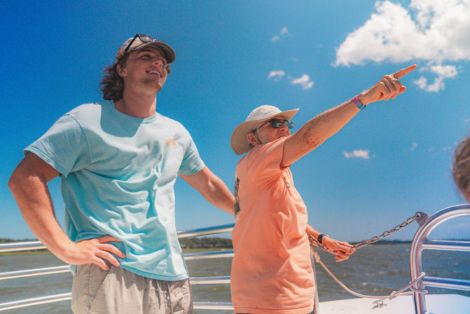 Two smiling men on a boat under a bright blue sky — a younger man in a light blue tee and a man in a sun hat and sunglasses pointing toward the coastal horizon over calm water.