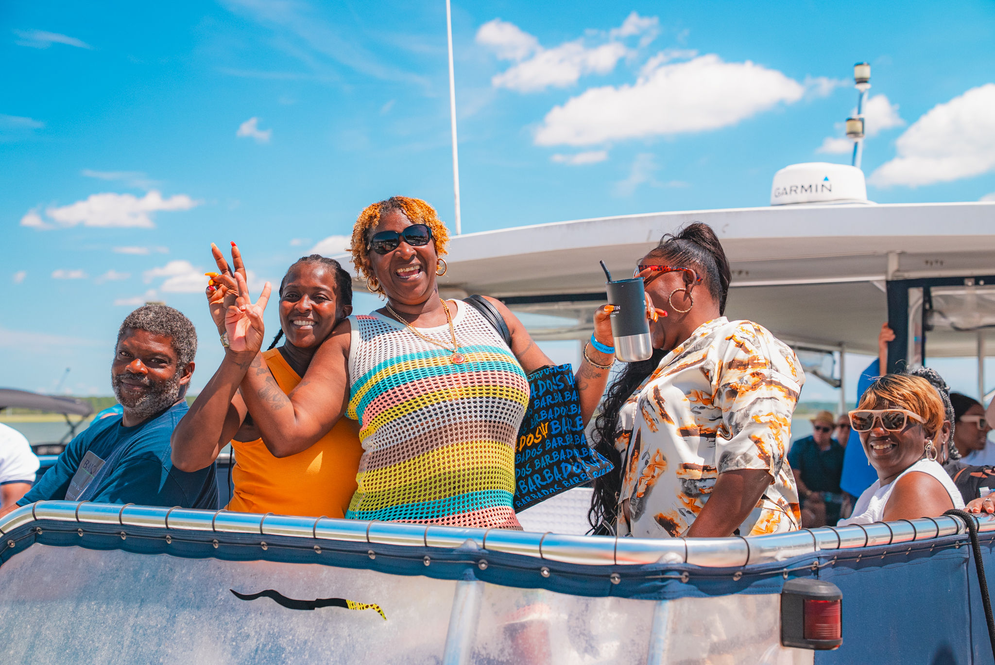 Smiling group of friends on a sunny boat cruise, wearing colorful summer outfits on the deck with blue sky and calm coastal water in the background