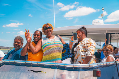 Smiling group of friends on a sunny boat cruise, wearing colorful summer outfits on the deck with blue sky and calm coastal water in the background