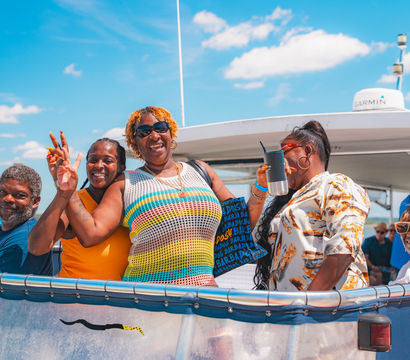 Smiling group of friends on a sunny boat cruise, wearing colorful summer outfits on the deck with blue sky and calm coastal water in the background