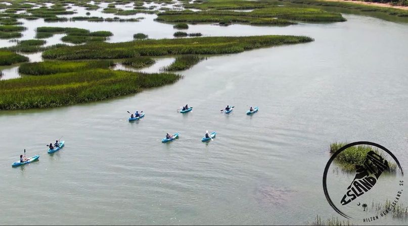 Aerial view of a group of kayakers paddling blue kayaks through winding tidal creeks and green salt marsh islands in a coastal wetland.