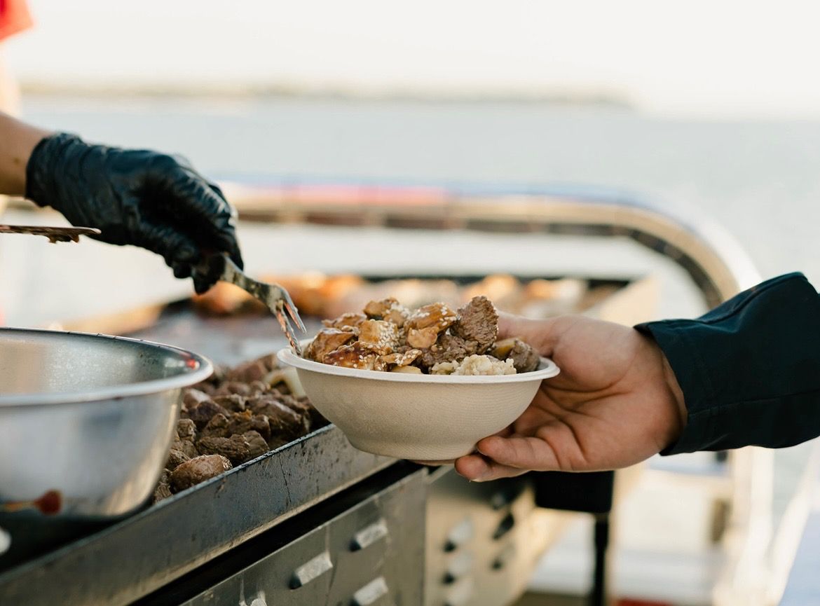 Waterfront street-food scene with a gloved server scooping grilled beef and rice into a to‑go bowl held by a customer at an outdoor barbecue.
