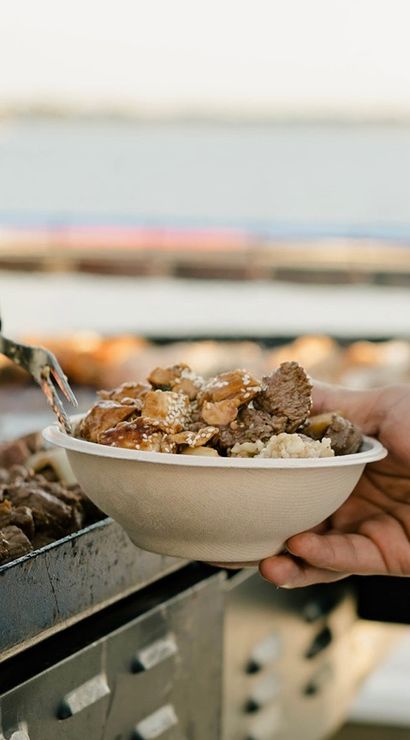Close-up of an outdoor food vendor serving grilled sesame beef over rice into a to-go bowl at a waterfront food stall.