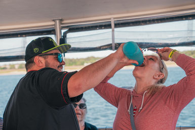 Man in a baseball cap and sunglasses holding a blue bottle as a woman in a pink hoodie tilts her head back to drink on a sunny boat, water and shoreline in the background.
