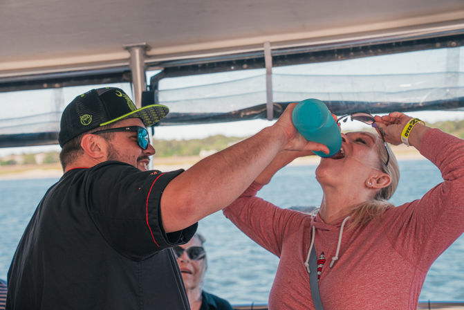 Man in a baseball cap and sunglasses holding a blue bottle as a woman in a pink hoodie tilts her head back to drink on a sunny boat, water and shoreline in the background.