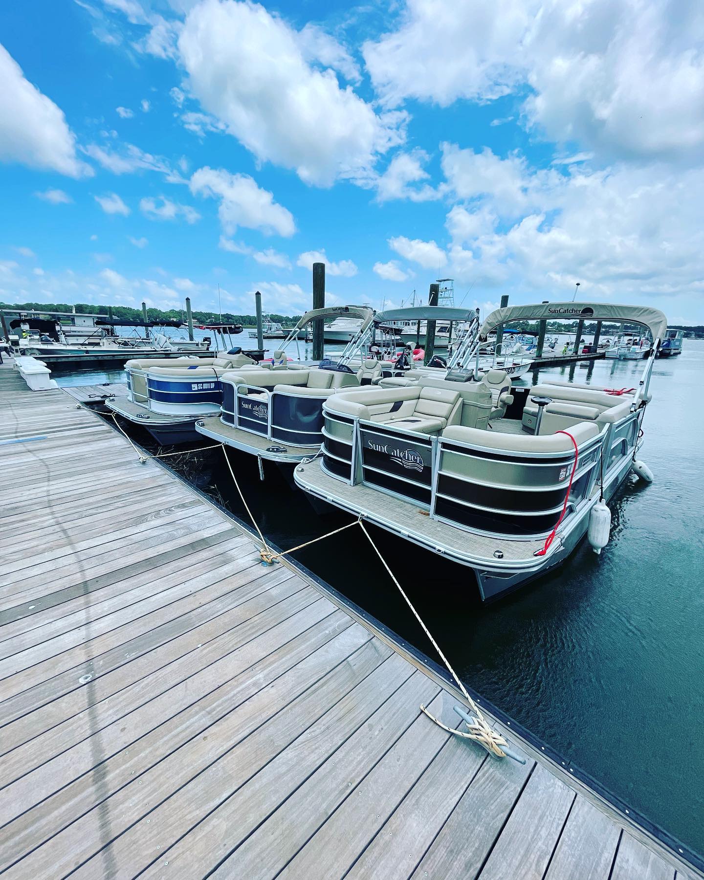 Three pontoon boats tied to a wooden marina dock on a calm lake under a bright blue sky with puffy clouds and other boats in the background