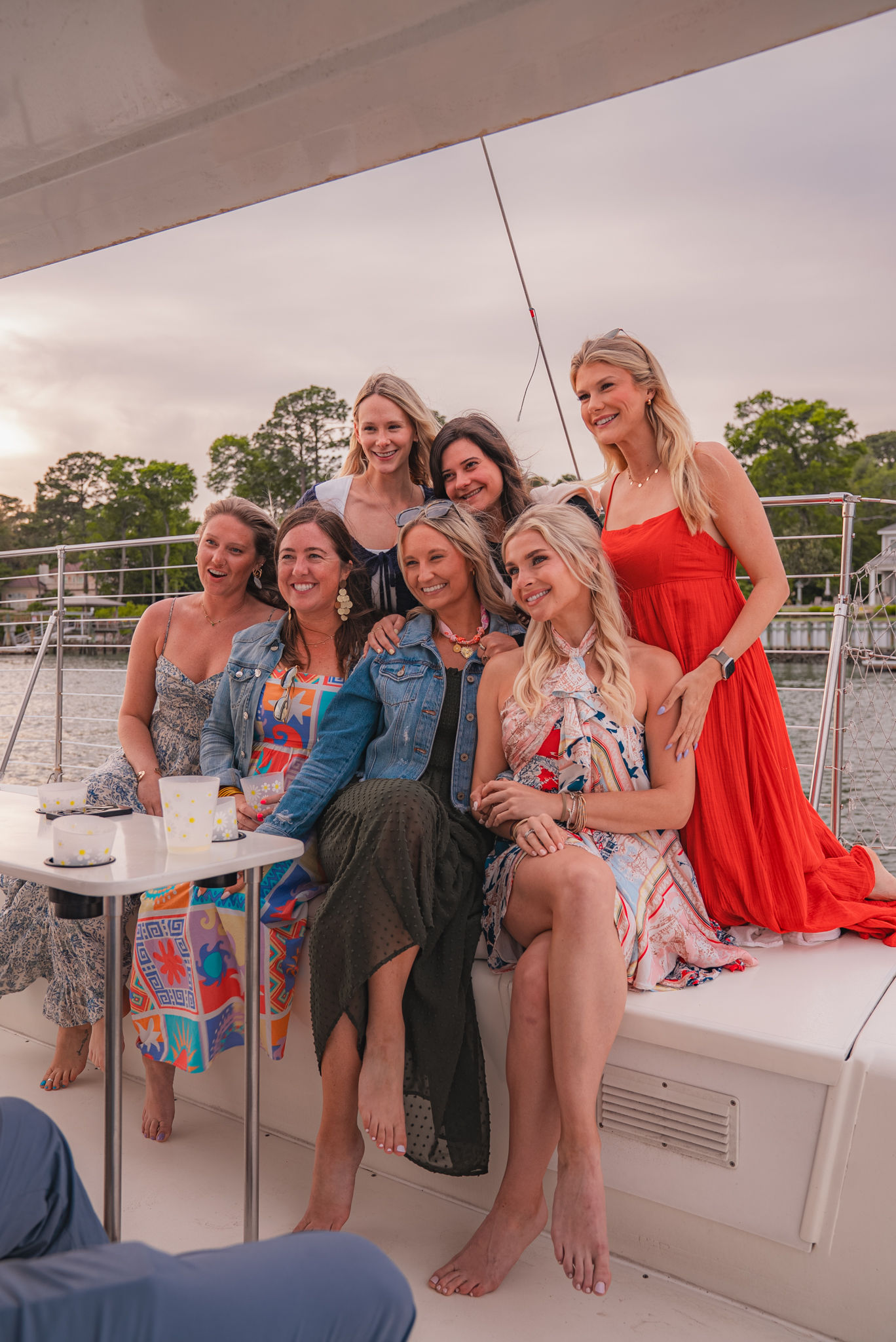 Seven women in colorful summer dresses smiling and posing barefoot on a sailboat deck during a sunset cruise, drinks on a small table and waterfront homes and trees visible in the background.