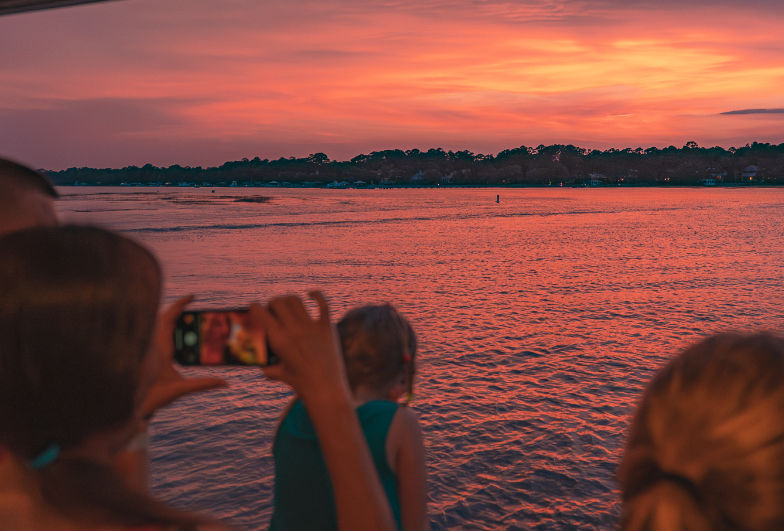 Group on a boat photographing a vibrant pink-orange sunset over calm water with a tree-lined horizon and glowing reflections.