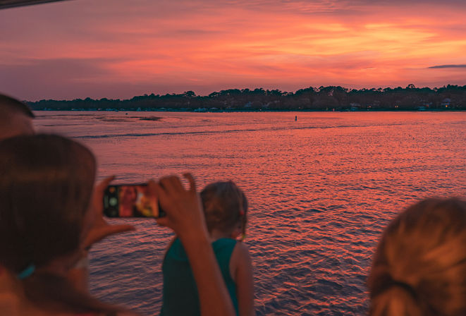 Group on a boat photographing a vibrant pink-orange sunset over calm water with a tree-lined horizon and glowing reflections.