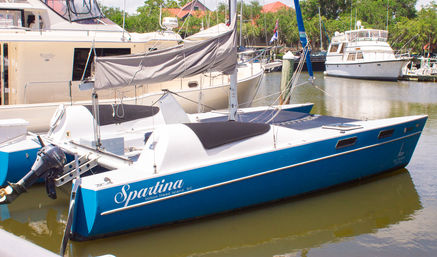 Blue catamaran sailboat docked in a sunny coastal South Carolina marina, folded gray sail and outboard motor visible, with larger yachts and green trees in the background.