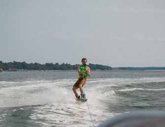 Wakeboarder in a bright green life vest towed behind a boat, cutting through spray on a calm coastal bay with a tree-lined shoreline and cloudy sky.