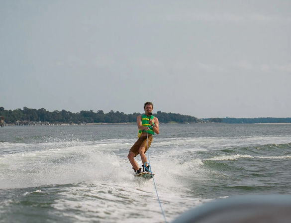 Wakeboarder in a bright green life vest towed behind a boat, cutting through spray on a calm coastal bay with a tree-lined shoreline and cloudy sky.