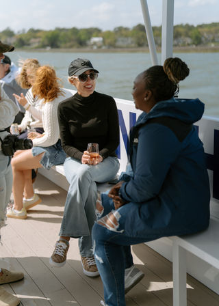 Two women chatting and smiling on a daytime coastal boat cruise — one wearing a black cap and sunglasses holding a wine glass, the other in a blue jacket with a braided bun, shoreline and calm water in the background.