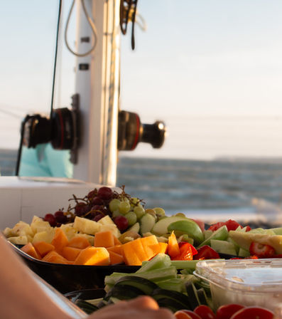 Bright fresh fruit platter (melon, grapes, strawberries, apples, cucumber) on a sailboat deck at sea with ocean waves and warm sunset light