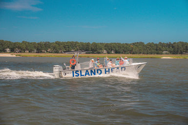 Tour boat with passengers cruising a coastal marsh inlet, cutting a white wake past a tree-lined shoreline under a bright blue sky