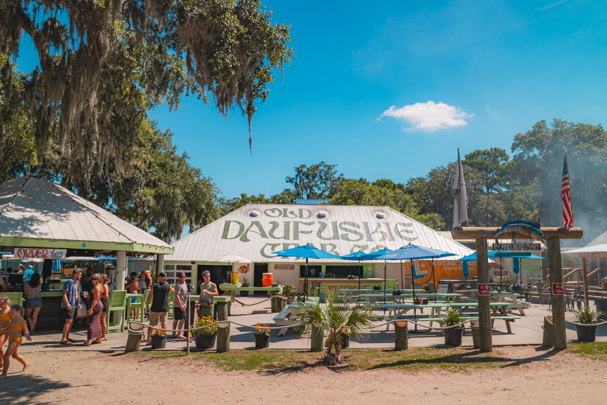 Sunny coastal outdoor seafood shack with picnic tables and blue umbrellas, oak trees draped in Spanish moss, casual summer crowds.