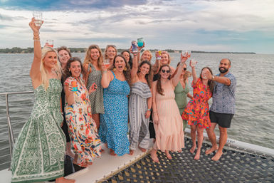 Lively group of adults cheering with drinks on the bow of a sailboat during a bay cruise, wearing colorful summer dresses and casual shirts against calm water and a cloudy sunset sky.