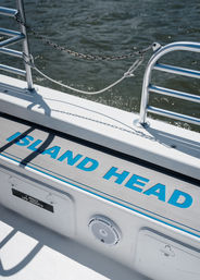 Sunlit boat deck step with bold turquoise lettering, stainless railings, chain and knotted rope casting shadows, small speaker and life-jacket locker visible, choppy water in the background.