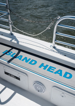 Sunlit boat deck step with bold turquoise lettering, stainless railings, chain and knotted rope casting shadows, small speaker and life-jacket locker visible, choppy water in the background.
