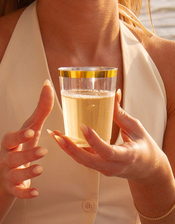 Close-up of a person with manicured nails holding a clear plastic cup with a gold rim filled with a bubbly drink, sunlit outdoor setting with warm sunset tones.