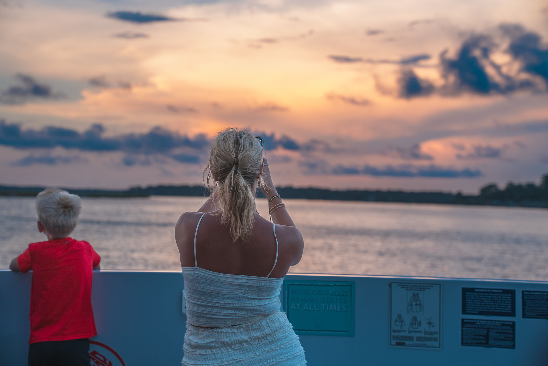 Woman capturing a pastel coastal sunset over calm water from a boat railing as a child in a red shirt watches the horizon.