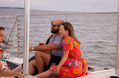 Couple relaxing on a sailboat deck with drinks during an ocean cruise — woman in a bright floral dress and man wearing sunglasses, calm water in the background.