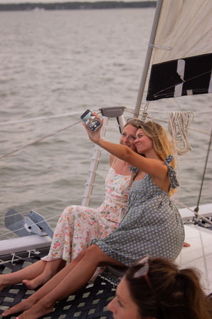 Two friends in summer dresses smiling and taking a selfie while relaxing on a sailboat’s deck net over calm water at sunset.