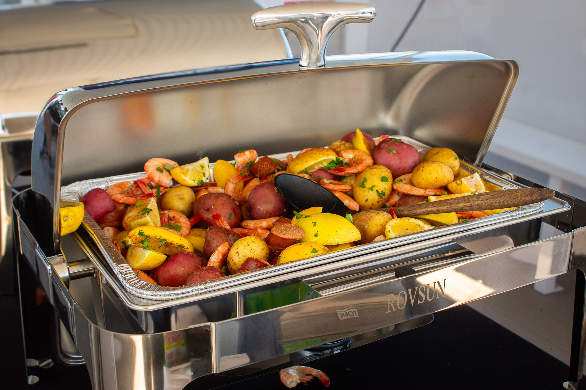 Stainless-steel buffet chafing dish filled with shrimp, red and yellow baby potatoes, sliced sausage, lemon wedges and parsley garnish — colorful hot seafood-style catering dish.