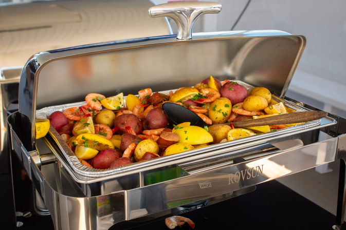 Stainless-steel buffet chafing dish filled with shrimp, red and yellow baby potatoes, sliced sausage, lemon wedges and parsley garnish — colorful hot seafood-style catering dish.