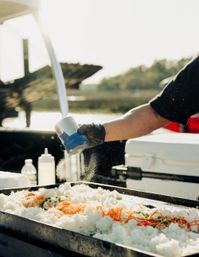 Gloved cook sprinkling seasoning onto a sunlit griddle of steamed rice and shredded carrots at an outdoor waterfront food stall