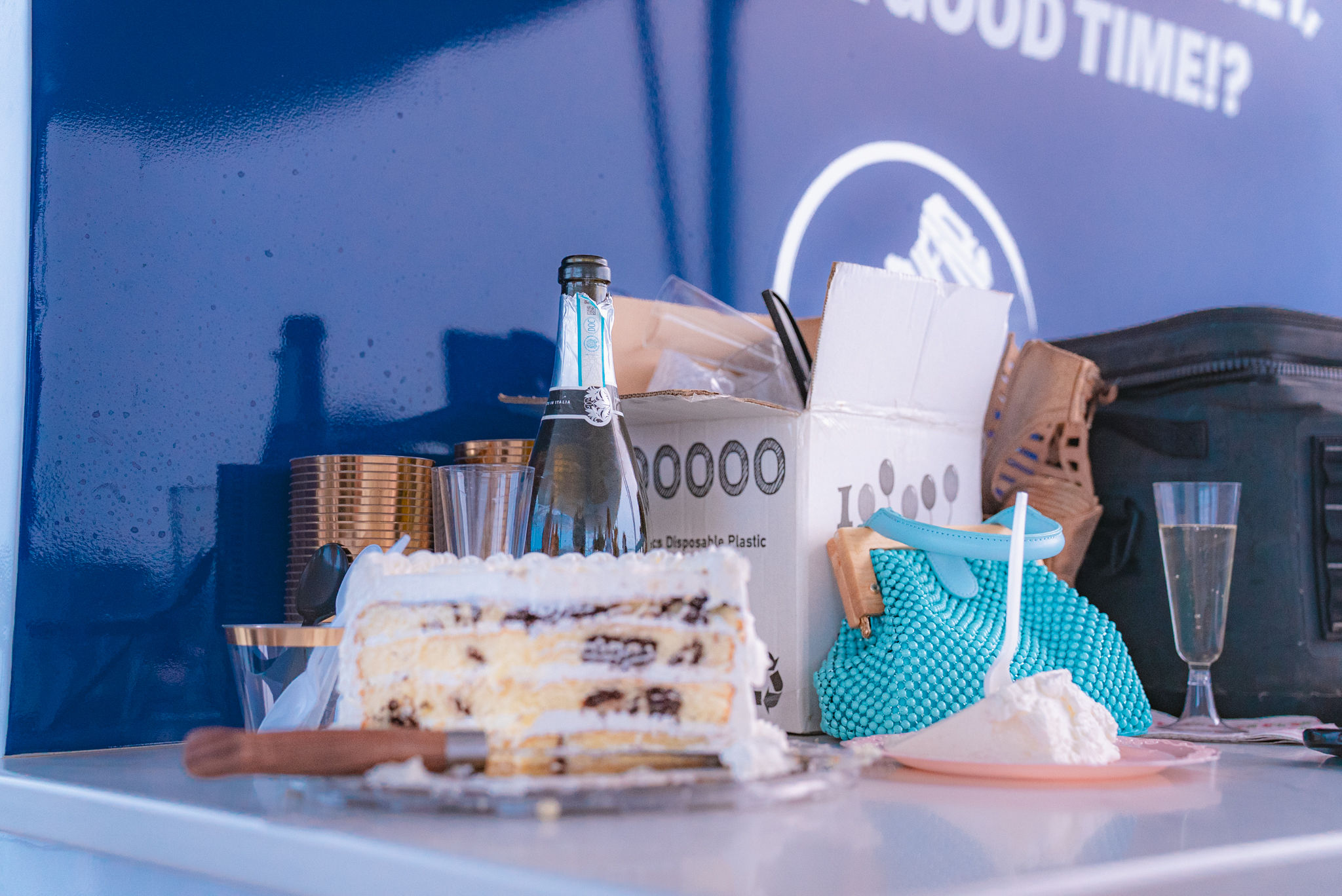 Party table with sliced layered cake, bottle of sparkling wine, plastic cups, a teal beaded purse and a cardboard box against a blue backdrop — casual celebration scene.