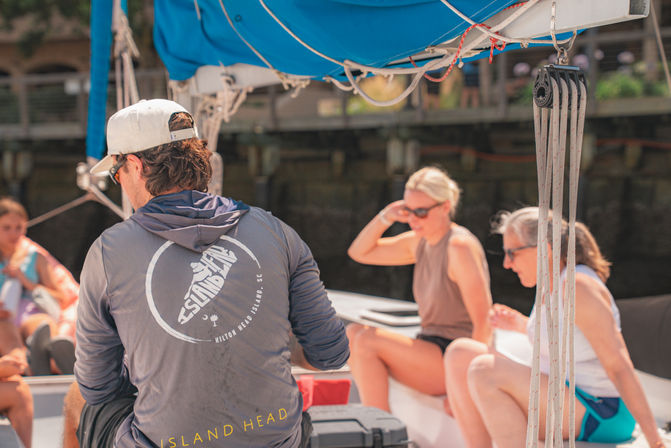 Leisurely group of adults relaxing on a sailboat near a marina at Hilton Head Island, SC, with a crew member in a cap and hoodie under a bright blue sail cover.