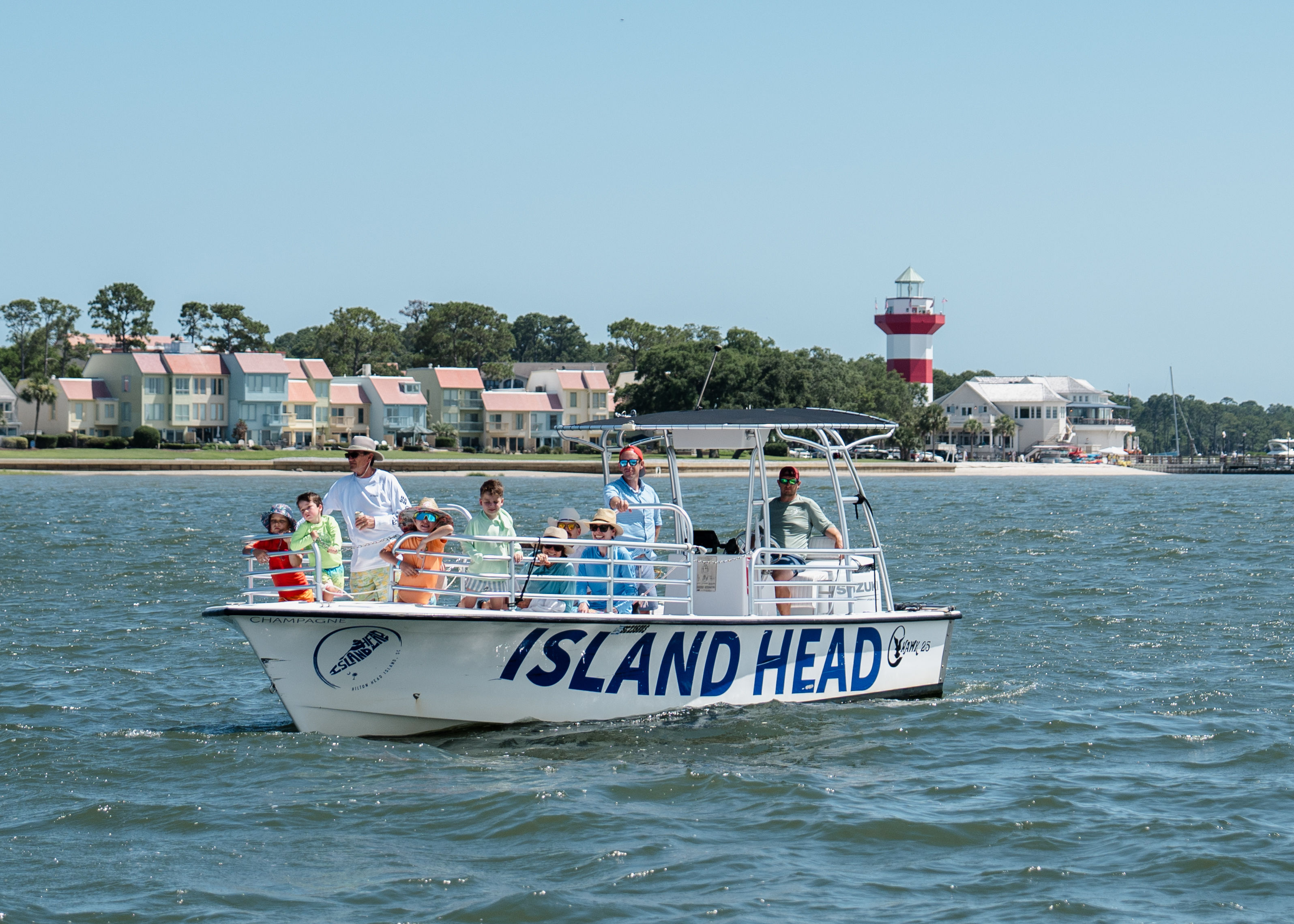 Family group on a white tour boat cruising a calm bay past pastel beachfront homes and a red-and-white lighthouse on a sunny coastal day