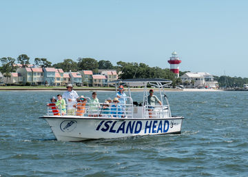 Family group on a white tour boat cruising a calm bay past pastel beachfront homes and a red-and-white lighthouse on a sunny coastal day