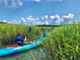 Kayakers paddling a bright turquoise kayak through a narrow coastal salt marsh channel flanked by tall green grasses under a sunny blue sky with fluffy clouds.
