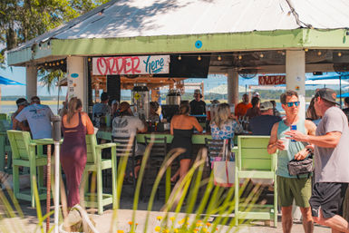 Sunny coastal waterfront tiki-style bar with green-painted counter and "Order Here" sign, patrons in summer clothes seated on high chairs and chatting under a metal roof with marsh and water in the background.