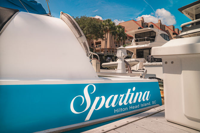 Turquoise-hulled boat docked at a sunny Hilton Head Island, SC marina with yachts and waterfront condos in the background.