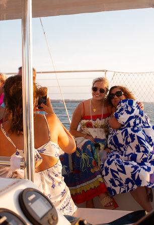 Three sunlit friends in colorful summer dresses posing and hugging for a phone photo on a sailboat at sunset with the ocean and horizon in the background.