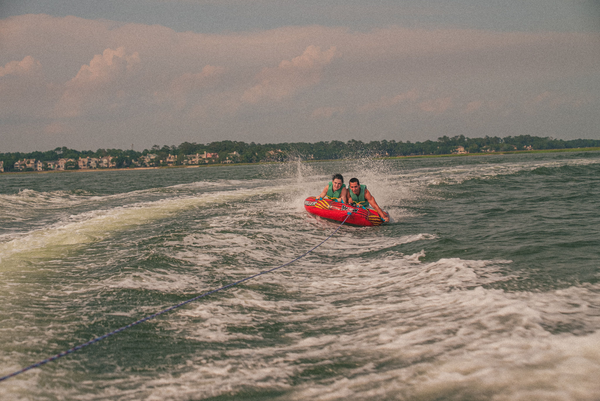 Two people in life jackets tubing on a red inflatable towable, splashing across a coastal bay behind a speedboat with a tree-lined shoreline and houses in the distance