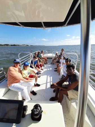 Cheerful group on a sunny pontoon boat cruise, seated along railings with calm blue coastal waters and a distant shoreline under a clear sky.