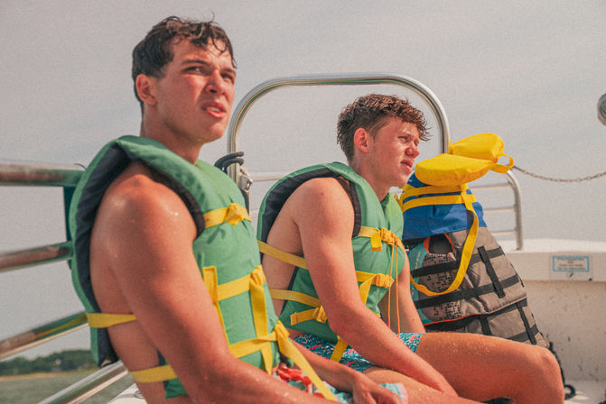 Two young men in green life jackets sit on a boat deck, squinting toward the sun with stacked colorful life vests and calm coastal water in the background — a fun summer boating scene.
