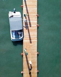 Aerial drone shot of a wooden dock with a moored motorboat, orange buoy fenders and long shadows over green marina water.
