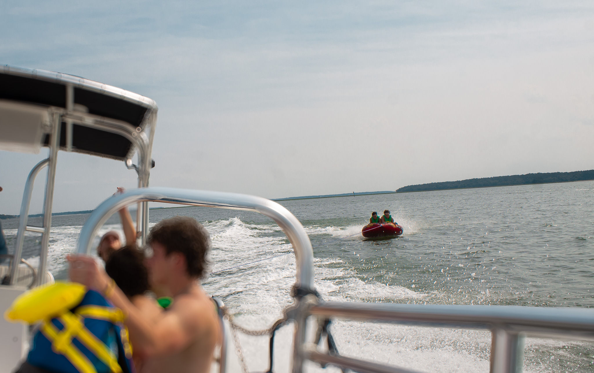 Summer tubing scene: two riders on a red inflatable towable pulled by a motorboat across a calm lake, seen from the boat deck with blurred passengers, metal railing and distant tree-lined shoreline.