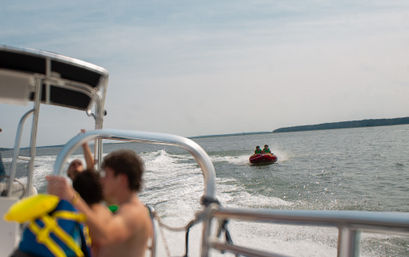 Summer tubing scene: two riders on a red inflatable towable pulled by a motorboat across a calm lake, seen from the boat deck with blurred passengers, metal railing and distant tree-lined shoreline.