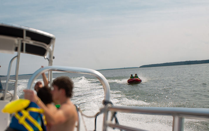 Summer tubing scene: two riders on a red inflatable towable pulled by a motorboat across a calm lake, seen from the boat deck with blurred passengers, metal railing and distant tree-lined shoreline.
