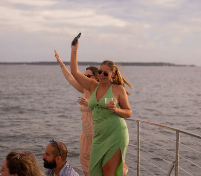 Two friends dancing and smiling on a boat deck during a coastal sunset cruise, woman in a green dress holding a drink and phone with ocean and distant shoreline in the background