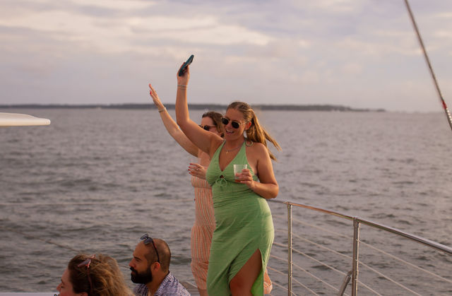 Two friends dancing and smiling on a boat deck during a coastal sunset cruise, woman in a green dress holding a drink and phone with ocean and distant shoreline in the background
