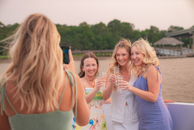 Three friends toast with champagne on a riverboat as a fourth takes their photo, tree-lined shore and dock visible at dusk