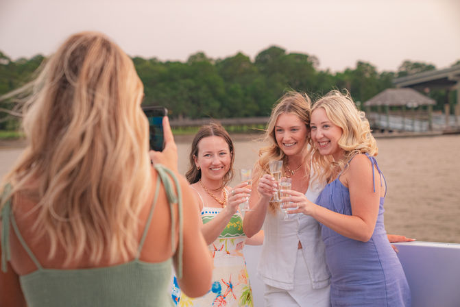 Three friends toast with champagne on a riverboat as a fourth takes their photo, tree-lined shore and dock visible at dusk